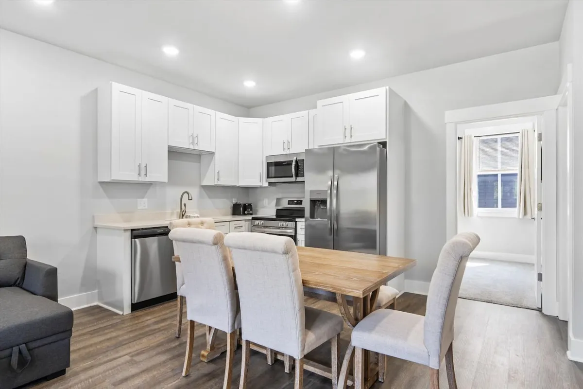 A close perspective on the dining table and kitchen appliances, with elegant lighting overhead. The setup emphasizes a cozy, functional space perfect for shared meals, cooking together, and long conversations over breakfast or dinner.