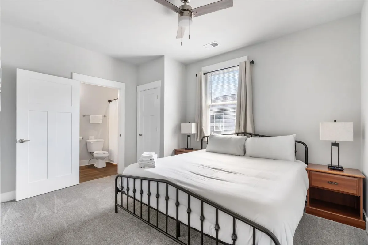 A tranquil bedroom featuring a king bed, soft linens, a nightstand, and a window letting in gentle sunlight. The doorway leads into the adjoining spaces, visually connecting the room to the rest of the upstairs area.