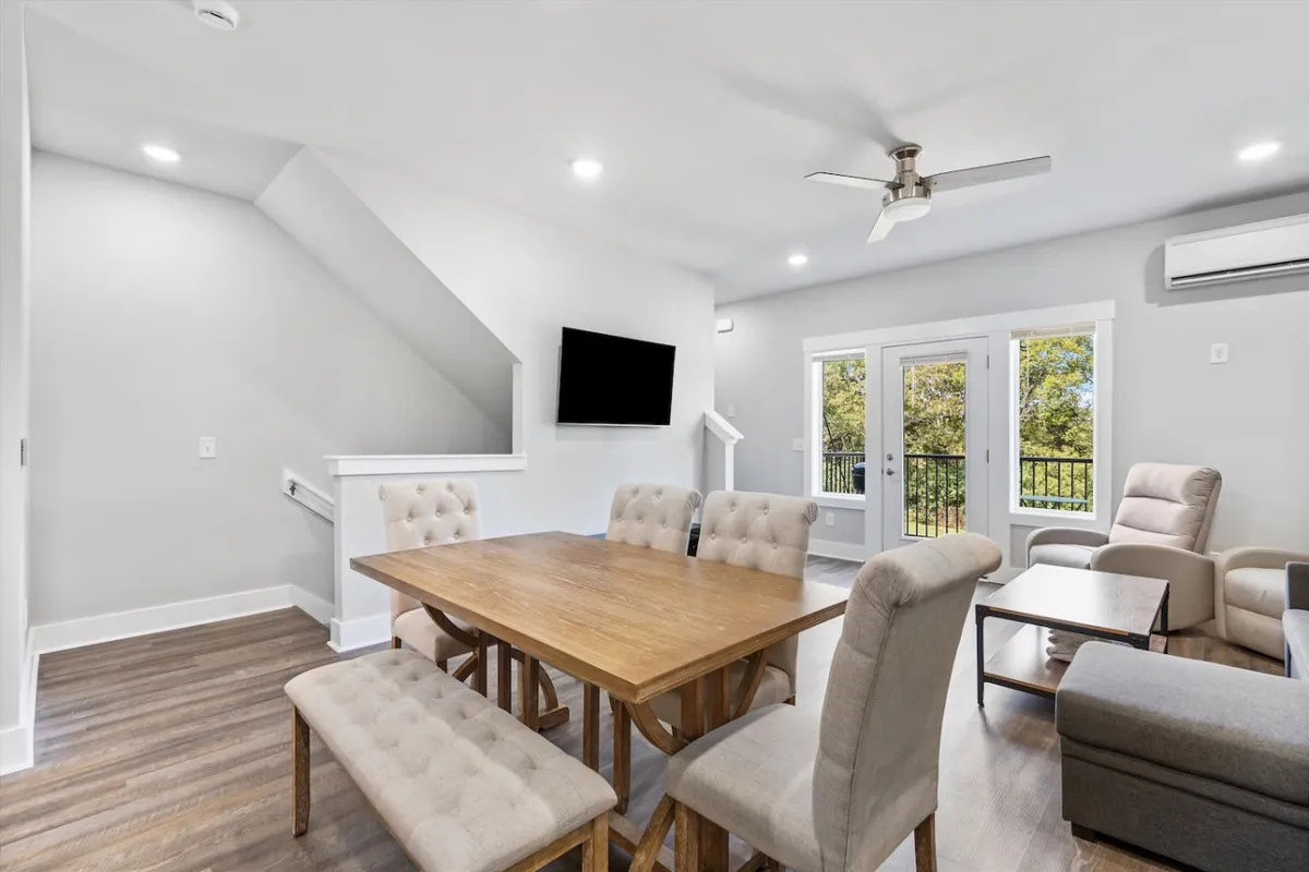 The image captures a bright, open-concept living and dining area with a spacious dining table set for gatherings, a comfortable sofa seating area facing a large TV, and expansive windows flooding the space with natural light. The scene feels cozy and inviting, perfect for group activities like dining, relaxing, or enjoying sunny mornings together.