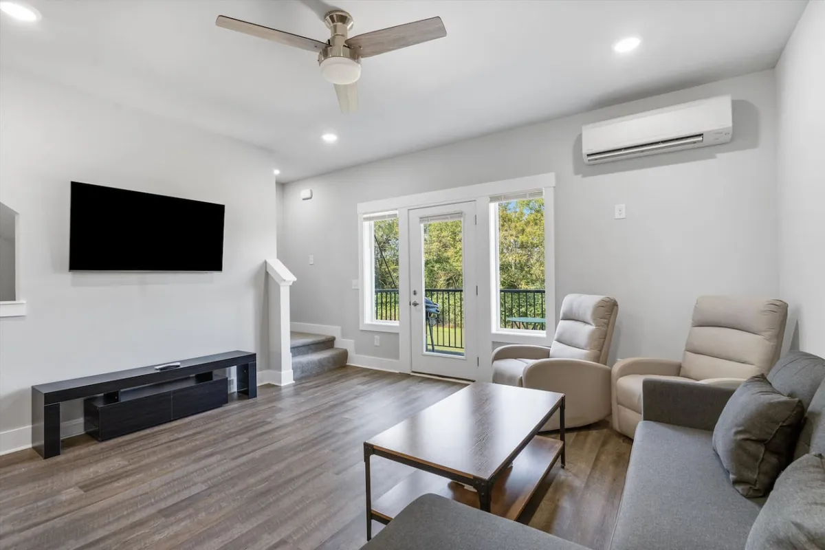 A cozy, comfortable living room featuring a sofa, coffee table, television, armchairs, and double doors that open to the outside. The space is arranged for both socializing and relaxing, with ample seating for a group.