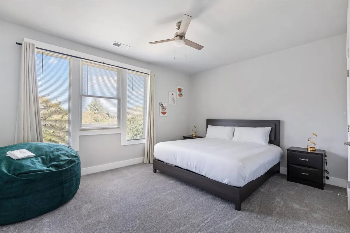 A serene bedroom with a plush king bed, bean bag chair by large windows, and abundant natural light streaming in, creating an inviting space for relaxing, reading, or enjoying a peaceful morning. The room features calming decor and soft linens, emphasizing comfort and tranquility.