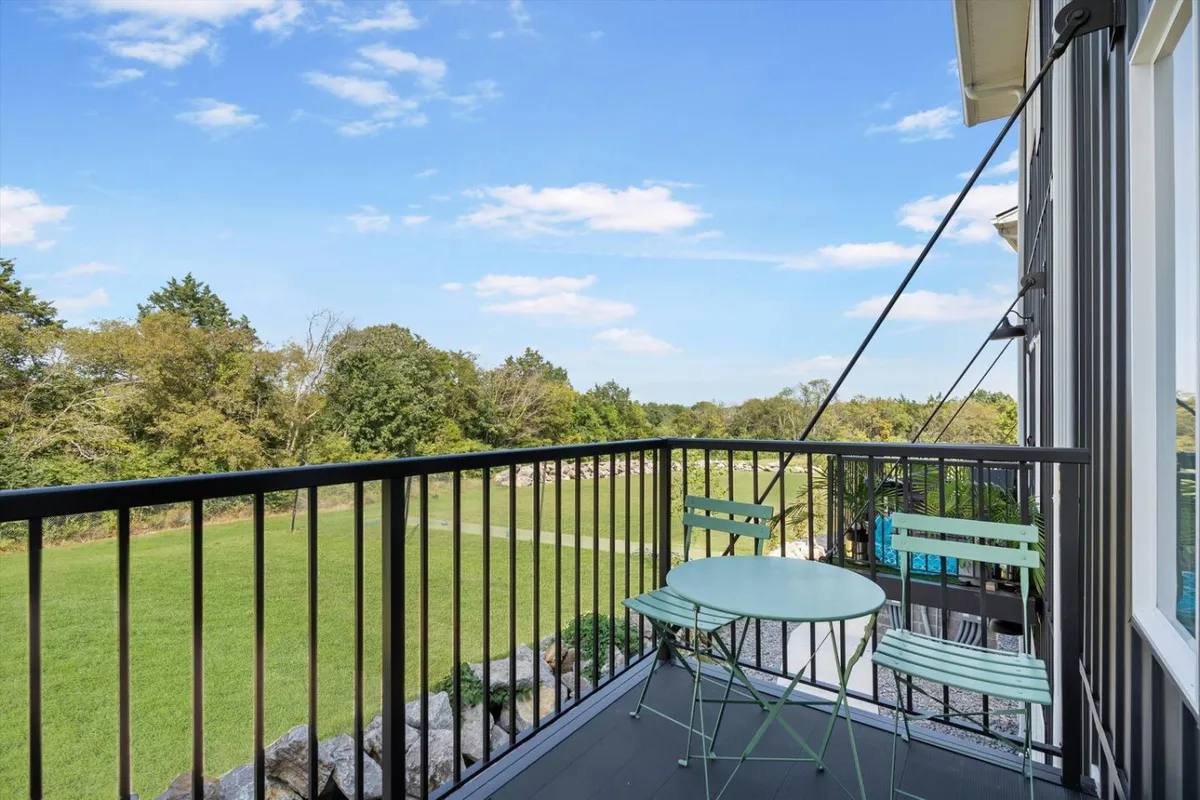 A welcoming exterior patio scene featuring a small round table with two green chairs set on an expansive green lawn, bordered by distant trees. The peaceful outdoor setting suggests a tranquil spot for morning coffee or taking work calls in fresh air.
