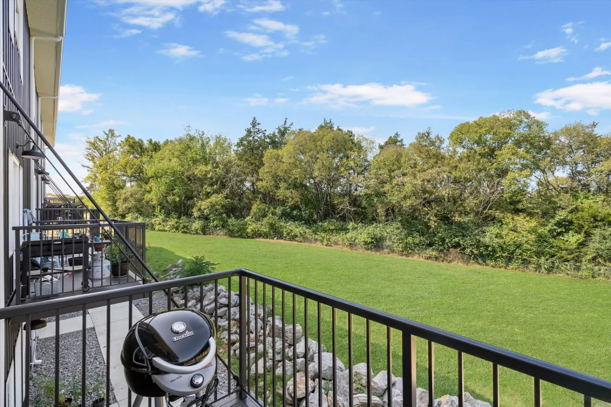 An inviting exterior scene featuring a balcony with a grill overlooking a well-maintained green lawn and a line of mature trees. The setting is perfect for barbecues, sunset views, or quiet mornings with coffee, blending the comforts of home with the outdoors.