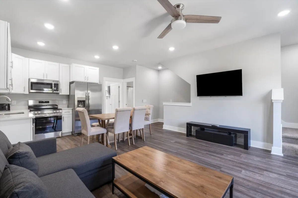 A wide view of an open-concept living area featuring a comfortable sofa, a spacious dining table, and a sleek kitchen area, all arranged to encourage conversation and group activities. The television is positioned for movie nights, while the decor and lighting create a warm, inviting ambiance.
